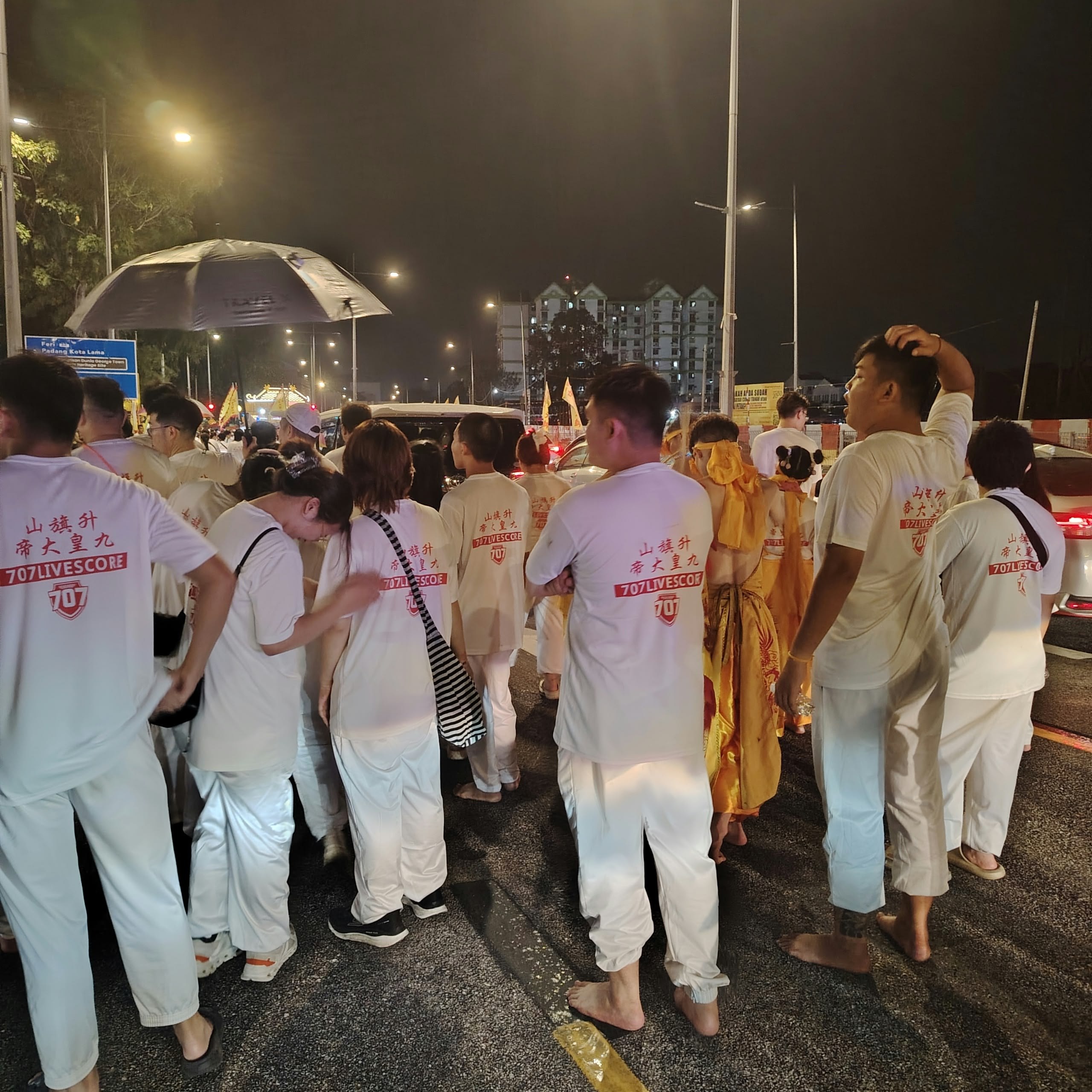 A nighttime street gathering with participants in white shirts bearing red 707Livescore branding, with flags and ceremonial activity visible.